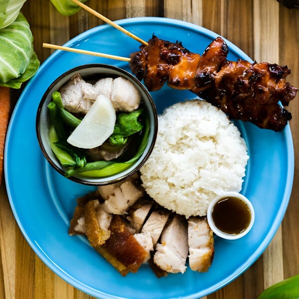 A blue plate with Filipino food: chicken skewers, rice, pork belly, and a vegetable dish.