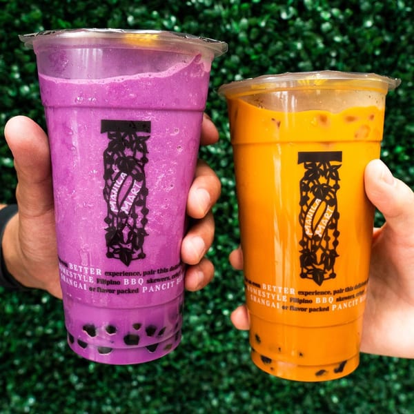 Hands holding two Manila Mart boba drinks, one purple ube and one orange, against a green background.