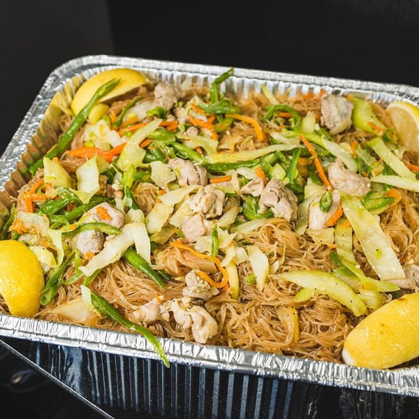 Filipino pancit bihon with chicken, vegetables, and lemon wedges in a foil tray.