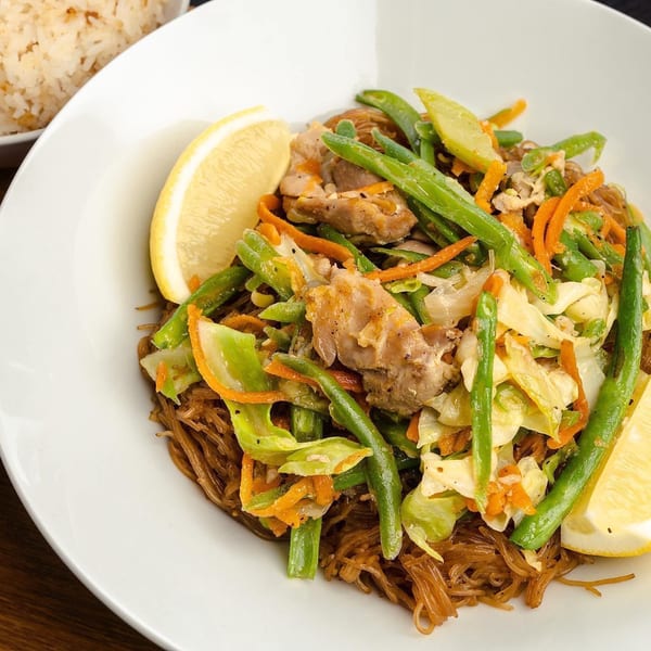 Filipino pancit bihon with chicken, green beans, carrots, cabbage, and lemon wedges, served with rice.